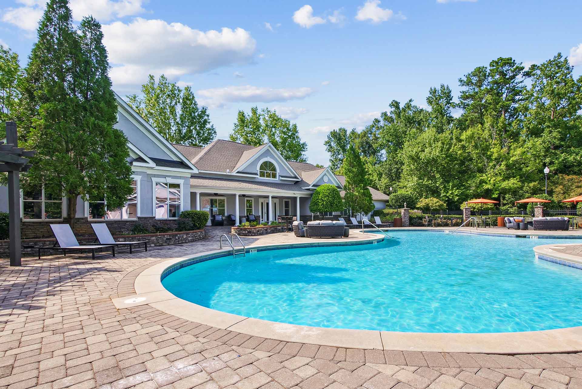 Halston South Point swimming pool with expansive sun deck, lounge chairs, tables with umbrellas and leasing office in background surrounded by native landscaping.