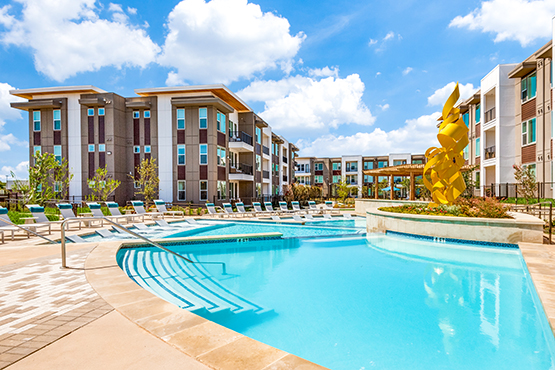 Pool with lounge chairs. A tall yellow sculpture in the flower bed and apartment buildings in the background.